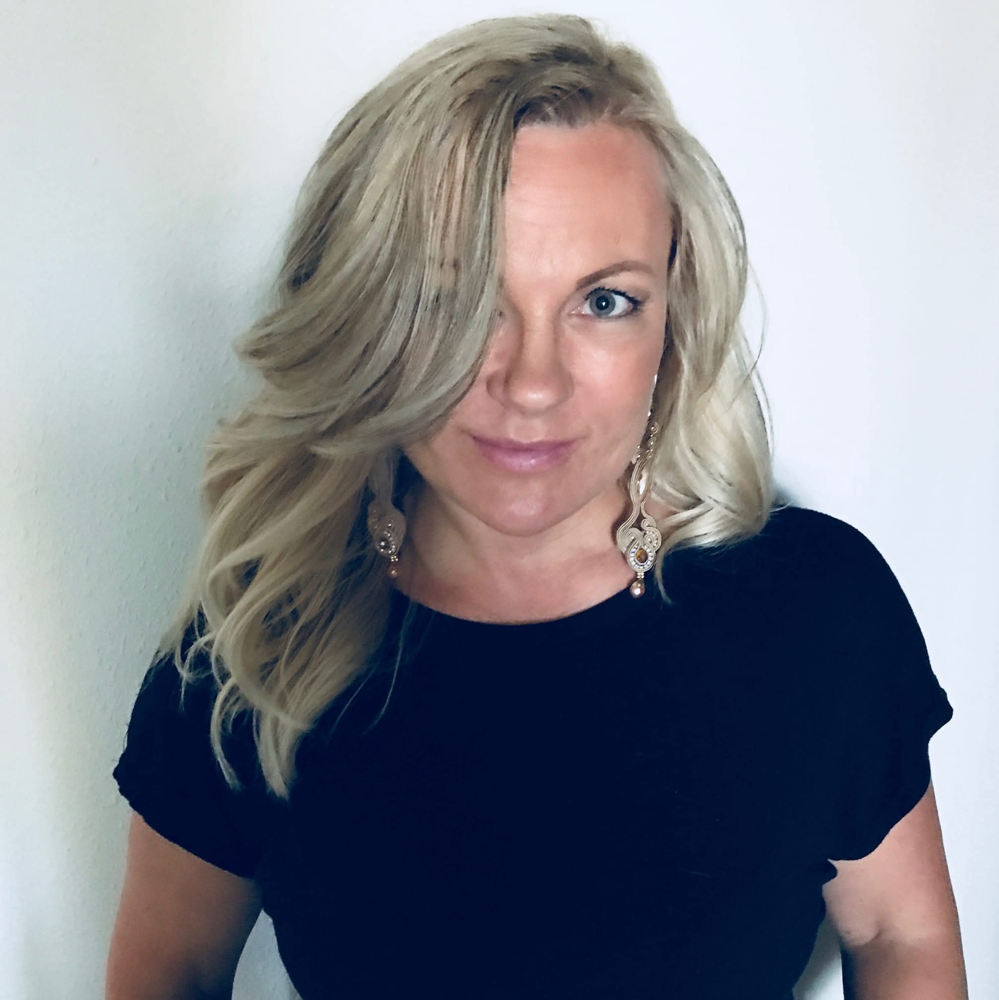Portrait of a smiling blonde woman posing against a light, neutral background. She wears a black blouse and decorative, dangling boho earrings. Her hair is styled in soft waves, and one section of her fringe slightly obscures one eye.