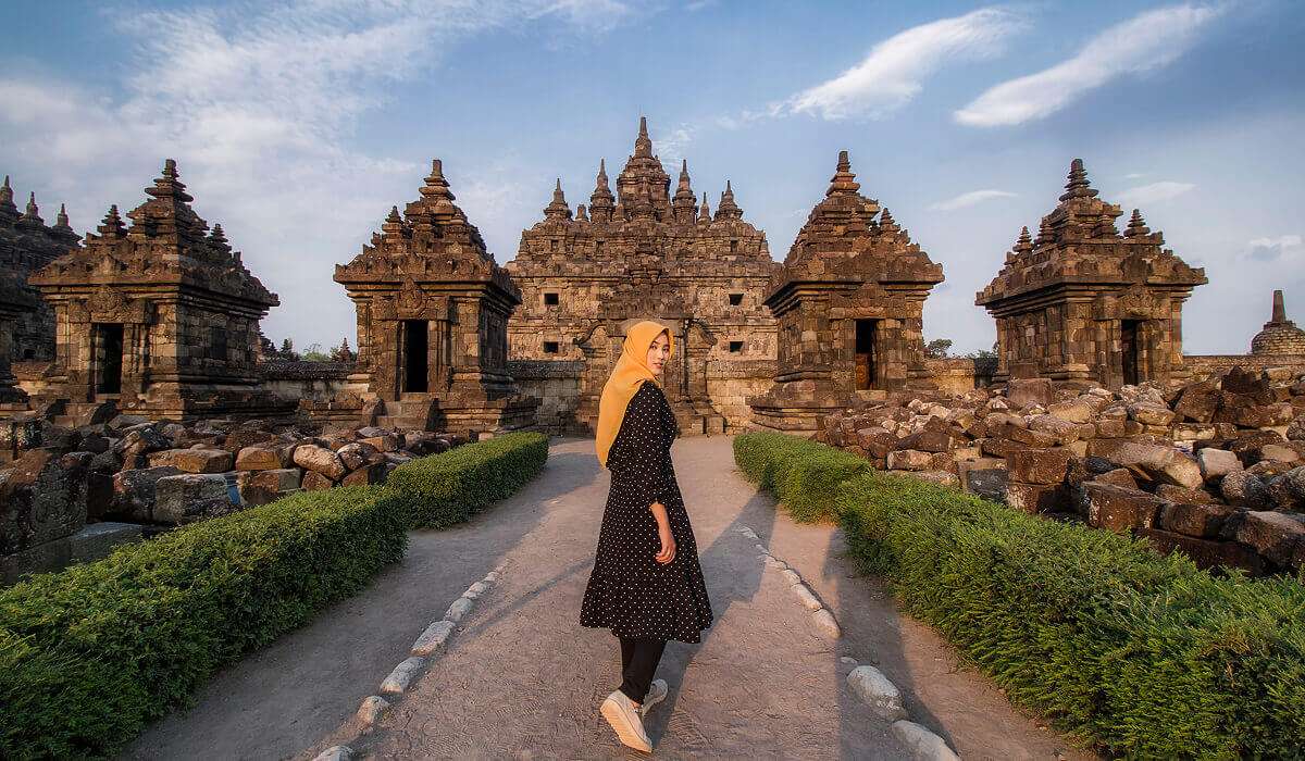 A woman in a yellow headscarf and dark dress stands with her back to the camera on the path leading to the ancient stone temple of Candi Plaosan. The structure is characterized by numerous turrets (stupas), and green hedges line the path.