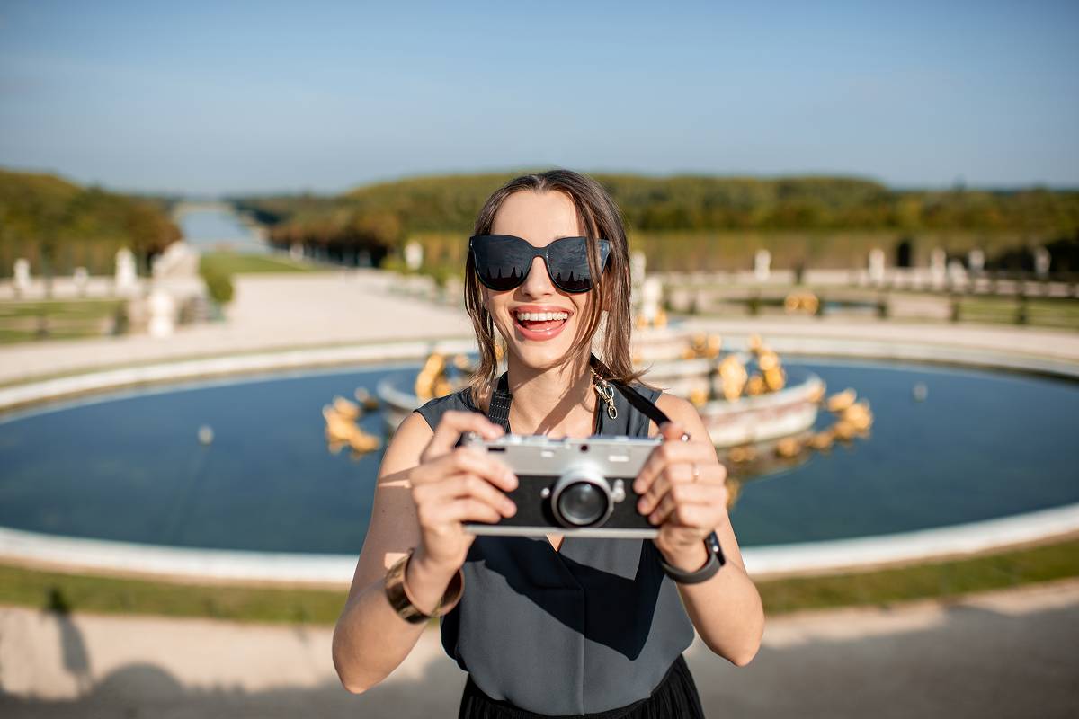 A close-up of a cheerful woman in sunglasses holding a camera with both hands, preparing to take a photo. In the background is a circular fountain pool and the extensive palace gardens.