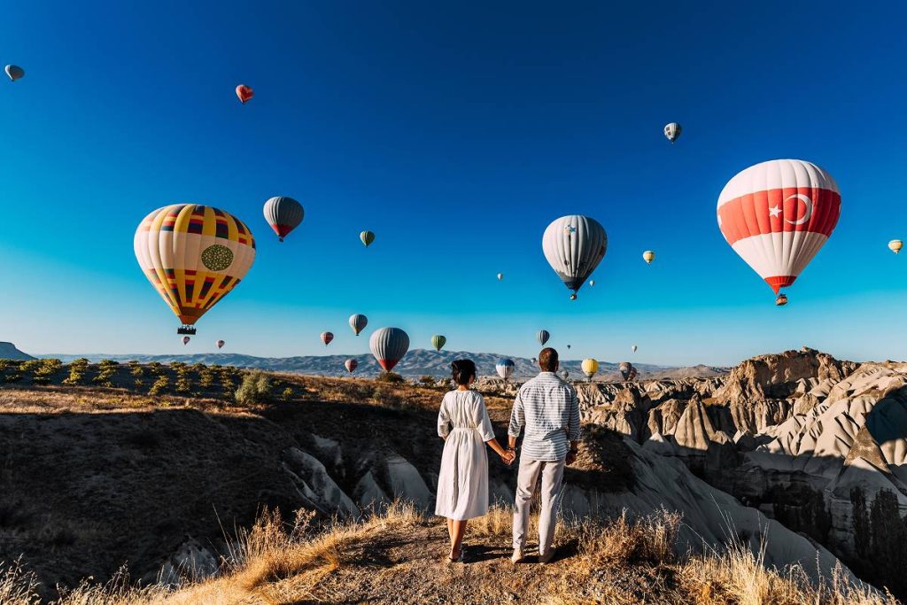 A couple holding hands stands on a cliff edge, looking out over a valley filled with dozens of colorful hot air balloons. The scenery features distinctive rock formations under a cloudless, deep blue sky.