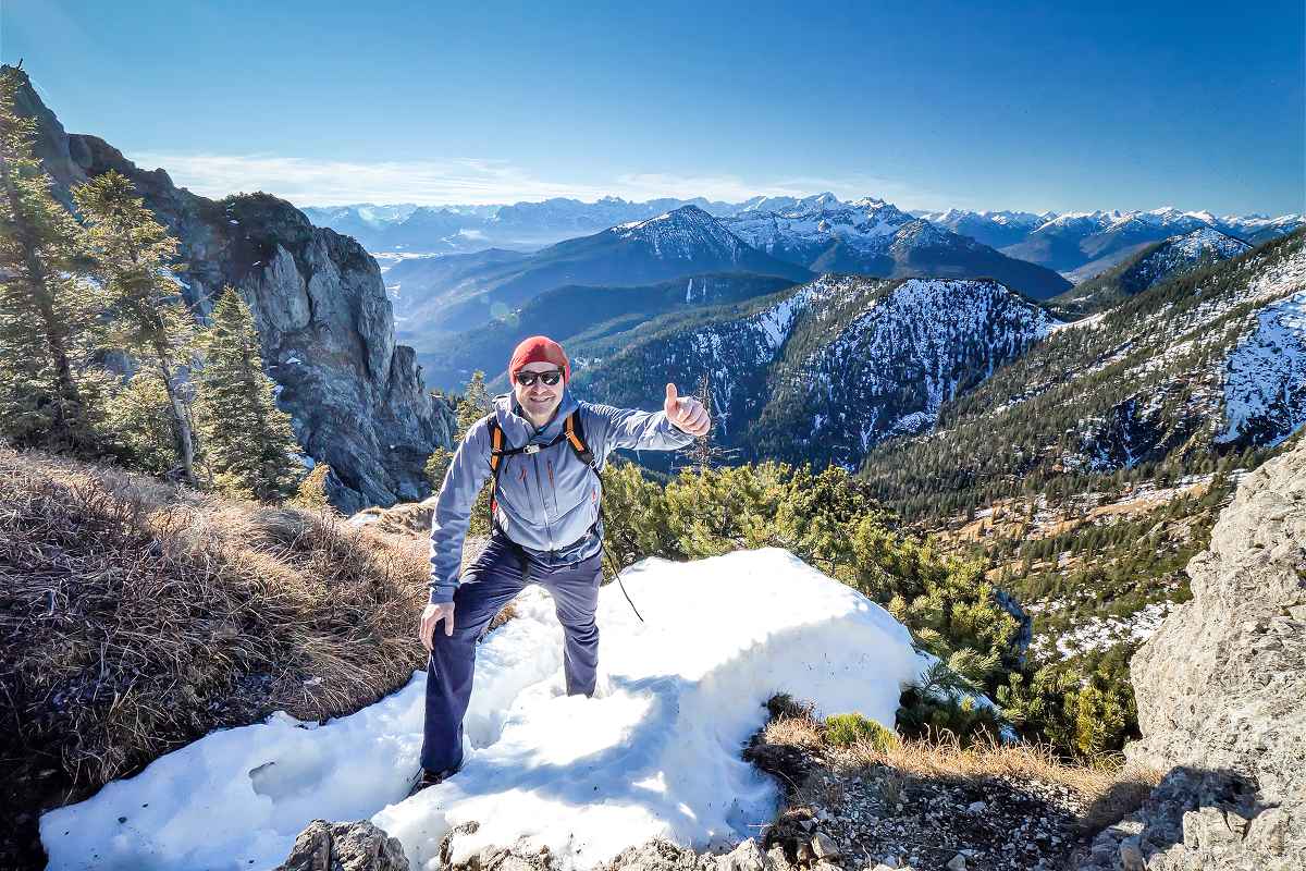 A man in a blue jacket, red hat, and sunglasses stands on a snowy mountainside. He gives the camera a thumbs-up sign, while behind him unfolds a sweeping panorama of forested valleys and distant snow-capped peaks under a clear sky.