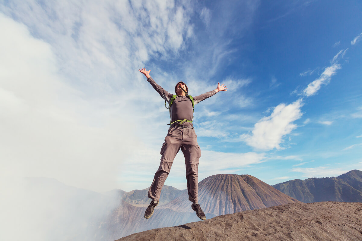 A man in trekking gear and carrying a backpack jumps high into the air against a blue sky dotted with white clouds. Volcanic mountain peaks are visible in the background, and the figure is perched on a sandy slope.