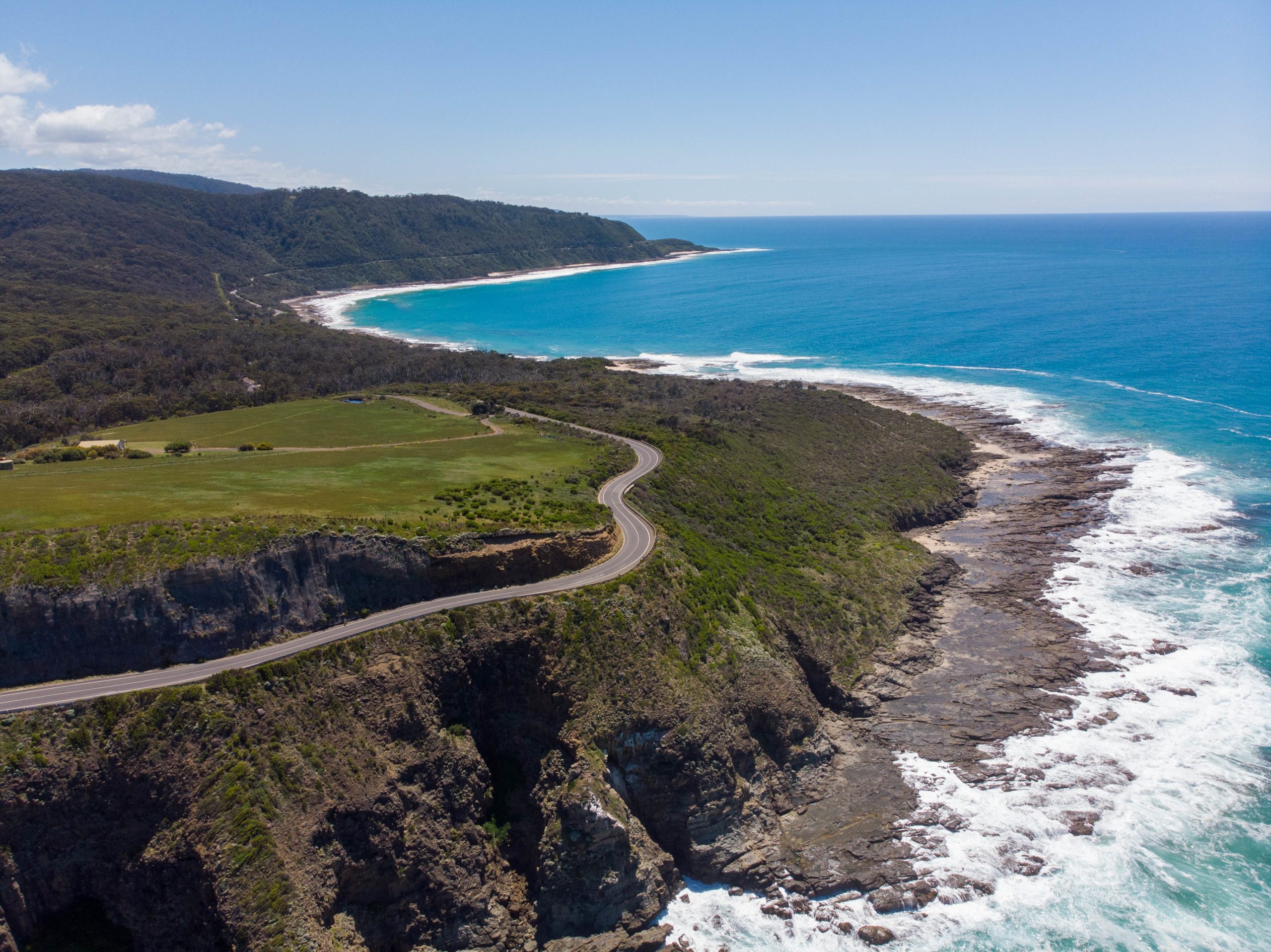 Aerial view of a winding asphalt road running along a high, green cliff above the ocean. On one side of the road, you can see dense forest and green glades, while on the other, foaming waves crash against the rocky shore and blue water.