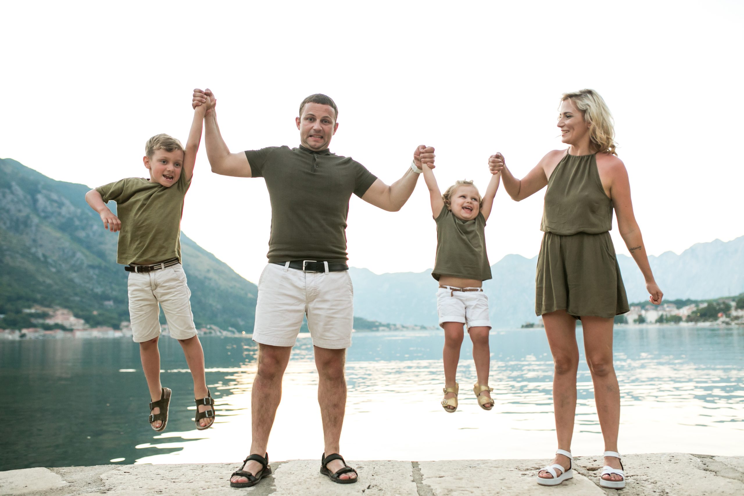 A joyful family of four sits on a quayside overlooking the water. The father and mother hold the hands of two children (a boy and a girl), lifting them up in a playful gesture. They are all dressed in olive green outfits and white shorts. In the background are mountains and a calm bay.